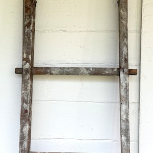 May include: A weathered wooden ladder with a rustic, aged appearance. The ladder has a simple design with vertical supports and horizontal rungs. The wood shows signs of wear, with peeling paint and visible grain. The ladder is leaning against a white wall.