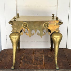 May include: A rectangular brass stand featuring a flat top surface with four small finials at the corners. The front panel displays two lion head motifs with attached rings, and the structure is supported by four curved legs.