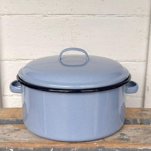 May include: A light blue enamel pot with a lid and two side handles. The pot has a black rim and a handle on the lid. The pot is sitting on a wooden surface.