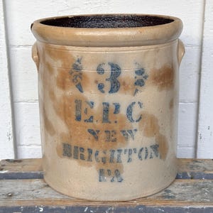 May include: Antique stoneware crock with a tan and brown finish. The crock features blue lettering that reads "3 E.P.C. NEW BRIGHTON PA". It has two small handles and a dark brown interior.