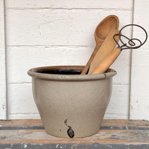 May include: A beige stoneware crock with a dark brown interior, holding wooden spoons and a wire whisk. The crock sits on a rustic wooden surface against a white-painted wood background. Kitchenware.
