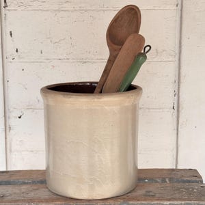 May include: A cylindrical ceramic crock with a smooth, light beige exterior and a dark brown interior. Inside the crock are three kitchen utensils: a large wooden spoon, a wooden spatula, and a smaller utensil with a green handle.