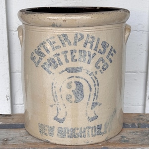 May include: A cylindrical beige stoneware crock featuring a dark blue stamped logo that reads ENTERPRISE POTTERY CO with a horseshoe graphic containing the number 3 and the text NEW BRIGHTON, PA at the base, including two side handles.