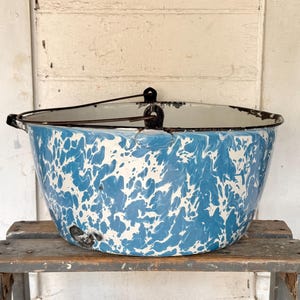 May include: A large, round, blue and white marbled enamel basin featuring a dark metal rim and a matching metal swing handle. The exterior displays a distinctive speckled pattern with visible signs of wear along the upper edge and base.