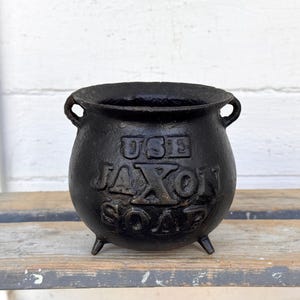 May include: A black cast iron cauldron with three small legs and two handles. The words "USE JAXON SOAP" are embossed on the front of the cauldron. The cauldron is sitting on a weathered wooden surface.