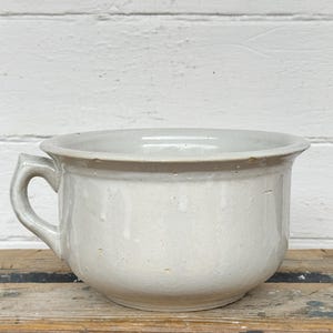 Könnte beinhalten: Cremefarbener Keramik-Nachttopf mit rundem Körper und kleinem Griff. Der antike Topf hat einen breiten Rand und steht auf einer Holzoberfläche vor einer weiß gestrichenen Wand. Ein Vintage-Stück für die Inneneinrichtung.