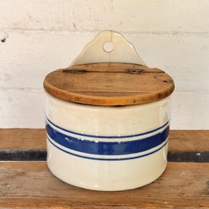 May include: A cylindrical off-white ceramic salt crock featuring a wide horizontal blue stripe around the center. The container has a hinged wooden lid and a raised back panel with a circular hole for wall mounting purposes.