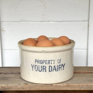 Puede incluir: Un cuenco de cerámica color crema lleno de huevos marrones. El cuenco tiene la inscripción "PROPERTY OF YOUR DAIRY" en azul. Los huevos son de color marrón natural. La escena está ambientada sobre una superficie de madera.