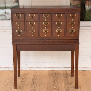 May include: A dark brown wooden cabinet featuring fifteen small drawers arranged in three rows of five. Each drawer is equipped with a brass label holder and a small brass pull handle. The cabinet is supported by four straight legs.