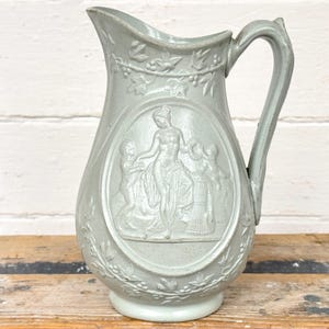 Op de afbeelding: Een lichtgrijze keramische kan met een handvat en een brede tuit. De kan heeft een reliëfontwerp van een klassieke scène met figuren en bloemmotieven. De kan is waarschijnlijk vintage en staat op een houten oppervlak.