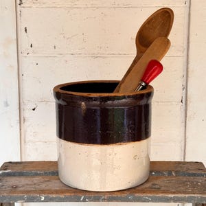 May include: A cylindrical stoneware crock featuring a two-tone design with a dark brown glazed upper section and a cream-colored lower section. Inside the crock are two wooden spoons and a kitchen tool with a bright red handle and metal shaft.
