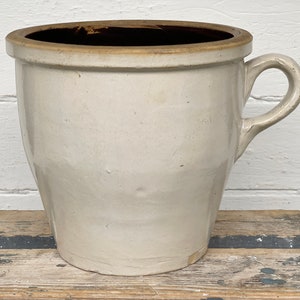 May include: A large, off-white stoneware crock with a handle. The crock has a wide, dark brown rim and a curved handle. The exterior is a matte, neutral color, and the piece appears to be vintage or antique.
