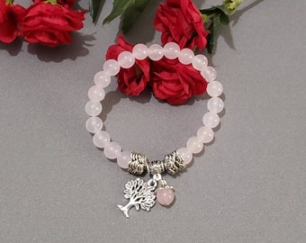 May include: A light pink beaded bracelet with a silver tree charm and a small pink bead. The bracelet is set against a gray background with red roses in the background.