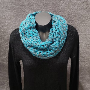 May include: A gray and turquoise crocheted infinity scarf. The scarf is worn on a black shirt.
