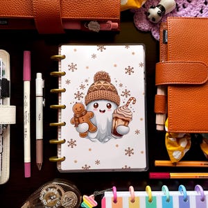May include: A collection of stationery items, including a planner with a winter-themed illustration of a ghost holding a gingerbread man and a coffee. Other items include pens, a tape dispenser, and a rainbow-striped notebook. The items are arranged on a dark surface.