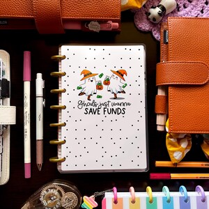 May include: A collection of stationery and planner accessories, including notebooks, pens, and decorative tape. A planner with the text "Ghouls just wanna SAVE FUNDS" is the focal point. The color palette includes orange, pink, white, and rainbow hues.