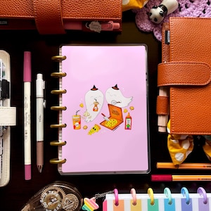 May include: A pink planner cover with a cartoon ghost design, pizza, and drink graphics. The image also includes brown leather planners, pens, washi tape, and a rainbow striped notebook.