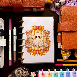 May include: An assortment of stationery items, including a planner with a ghost illustration, pens, and decorative tape. The ghost design features a cute cartoon ghost with orange bows and floral accents. Other items include orange leather planners and colorful paper clips.