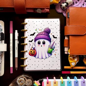 May include: A collection of stationery items, including a planner with a Halloween ghost illustration, pens, and decorative accessories. The planner features a cute ghost character with a pumpkin bag and a drink. Other items include a rainbow striped notebook and leather-bound journals.