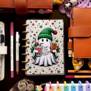 May include: A collection of stationery items, including notebooks, pens, and decorative accessories. A central notebook features a ghost illustration with the text "Don't Stop Believin'". The color palette includes orange, white, green, and pink.