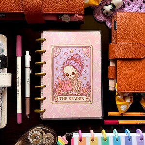 May include: A collection of stationery items, including notebooks, pens, and decorative accessories. A planner with a pink cover featuring a skeleton reading a book is the focal point. Other items include a rainbow striped notebook, orange leather planners, and colorful pens.