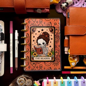 May include: An assortment of stationery items, including orange and white planners, pens, and decorative accessories. A planner with a skeleton reading a book is the focal point, with the text "THE READER". Other items include a rainbow striped notebook and decorative bows.
