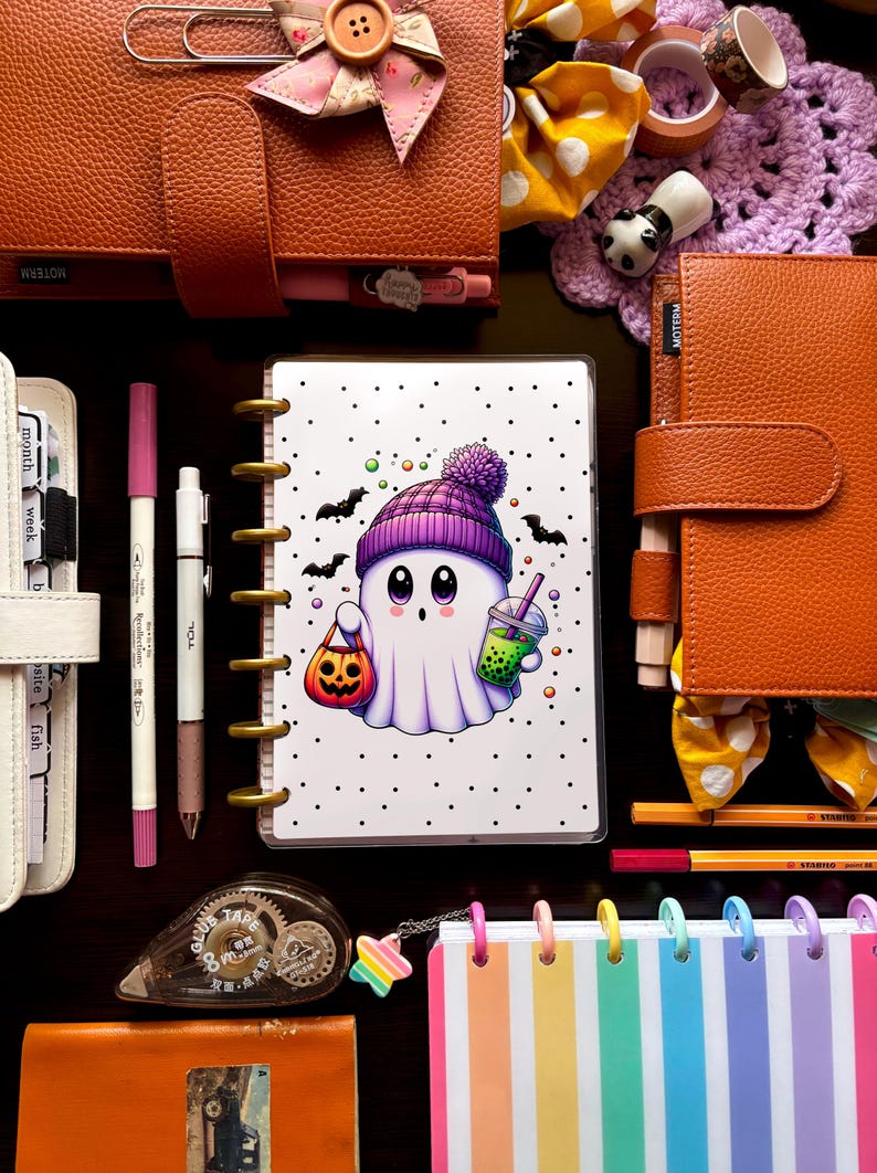 May include: A collection of stationery items, including a planner with a Halloween ghost illustration, pens, and decorative accessories. The planner features a cute ghost character with a pumpkin bag and a drink. Other items include a rainbow striped notebook and leather-bound journals.