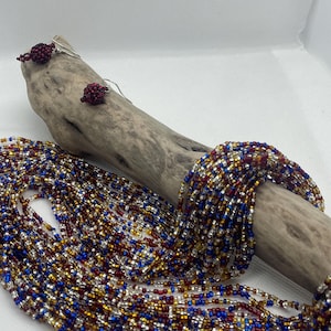 Op de afbeelding: Een meerstrengs kralenketting en bijpassende oorbellen. De ketting heeft strengen kleine kralen in rood, blauw, goud en zilver. De oorbellen zijn kleine, ronde clusters van rode kralen. De sieraden worden tentoongesteld op een stuk drijfhout.