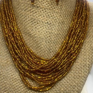 May include: A gold beaded necklace and matching earrings set. The necklace features multiple strands of small, shiny gold beads. The earrings are also gold and beaded, with a round design. The jewelry is displayed on a burlap-covered bust.