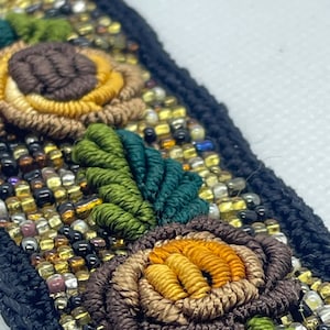 May include: Close-up of a black embroidered textile with floral and leaf designs. The flowers are brown and yellow, with green leaves. The textile is embellished with gold, brown, and clear beads.