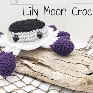 May include: A crocheted crab toy with purple, white, gray, and black yarn. The crab is sitting on a piece of driftwood.
