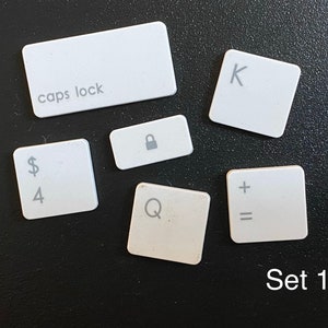 May include: A collection of white keyboard keys, including "caps lock", "K", "Q", "+", "=", a lock symbol, and a key with "$4". The text "Set 1" is also visible. The keys are arranged on a dark surface.