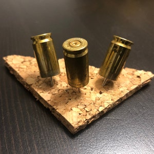May include: Three brass bullet casings repurposed as push pins, mounted on a small, irregularly shaped piece of cork. The casings are a warm gold color, and the cork is a light brown. The pins are arranged in a triangular pattern.