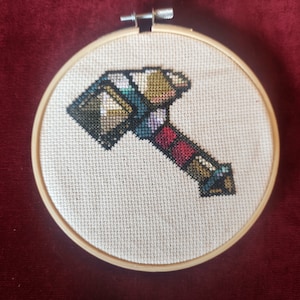 Peut inclure: Un cerceau de broderie au point de croix avec une image pixélisée d'un marteau dans différentes nuances de brun, rouge, bleu et vert.