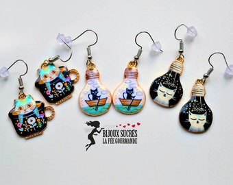 May include: A set of six cat-themed earrings. Two earrings feature a black cat in a boat inside a lightbulb. Two earrings feature a white cat inside a lightbulb with a starry sky. Two earrings feature a white cat inside a black teacup with flowers.