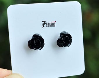 May include: A pair of black rose-shaped earrings on a white card. The earrings are small and detailed, with visible petals. The card also features the text "BIJOUX SUCRÉS LA FÉE GOURMANDE" and a small graphic. The background is a blurred green.