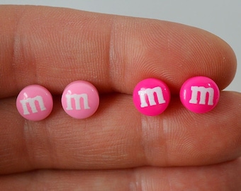 May include: Four pink and white M&M candy-shaped earrings. The earrings are displayed on a hand.