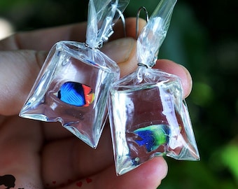 May include: Unique earrings featuring miniature colorful fish inside clear plastic bags. The fish have vibrant blue, red, and yellow hues. The earrings are suspended from silver-colored hooks, creating a whimsical accessory.