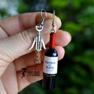 May include: A pair of silver earrings, one shaped like a corkscrew and the other shaped like a bottle of Casillero del Diablo Carmenere wine. The bottle is red and black with a white label.