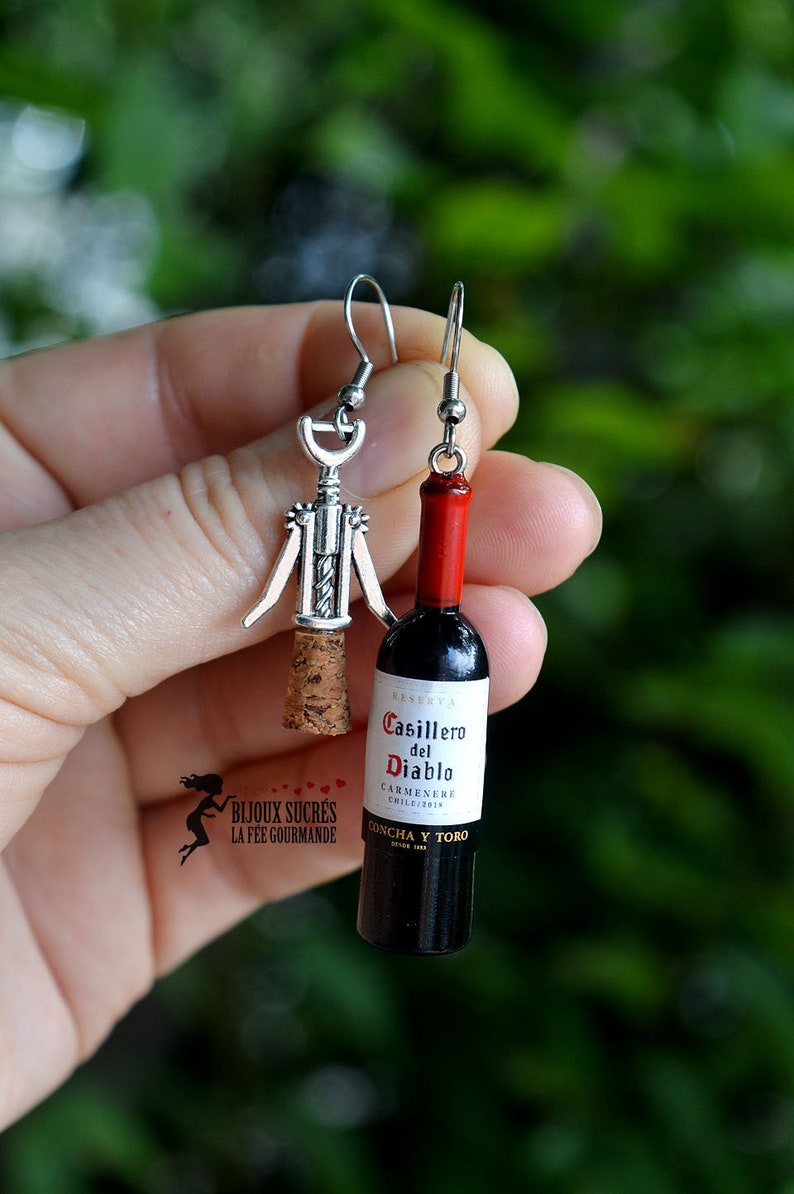 May include: A pair of silver earrings, one shaped like a corkscrew and the other shaped like a bottle of Casillero del Diablo Carmenere wine. The bottle is red and black with a white label.