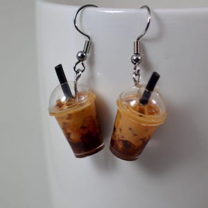 May include: A pair of dangle earrings shaped like miniature iced coffee drinks. Each earring has a clear plastic cup with a brown and tan liquid, a black straw, and a silver-tone fish hook.