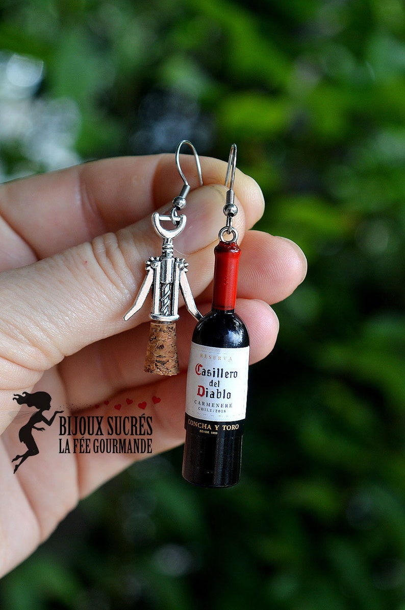 May include: A pair of silver earrings featuring a miniature corkscrew and a miniature bottle of red wine. The wine bottle label reads "Casillero del Diablo Carmenere Chile 2019 Concha y Toro".