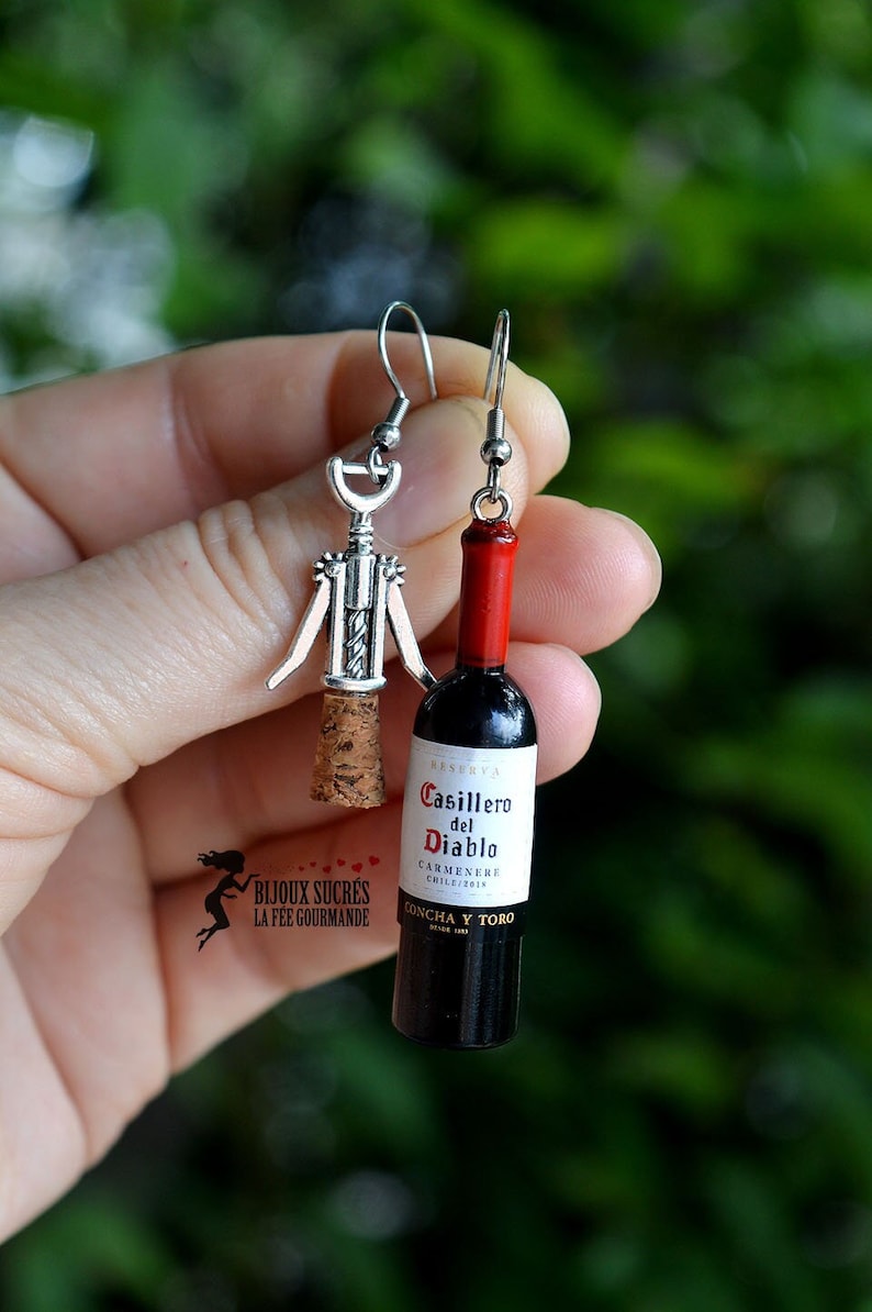 May include: A pair of silver earrings featuring a miniature wine bottle and a corkscrew. The wine bottle is labeled "Casillero del Diablo Carmenere Chile/2014 Concha y Toro".