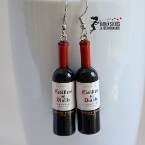 May include: A pair of earrings featuring miniature wine bottles with the label "Casillero del Diablo Carmenere Chile/2015 Concha y Toro" in black and white text on a red and black bottle.