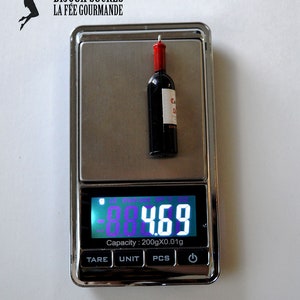 May include: A small, red wine bottle charm weighs 4.7 grams on a digital scale. The charm is labeled "C.D. Co."