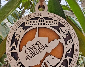 May include: A wooden ornament shaped like the state of West Virginia with the words "West Virginia" inside the state outline. The ornament is decorated with various icons representing different locations in West Virginia, including Seneca Caverns, Harpers Ferry, Berkeley Springs, and the New River Gorge National River. The ornament is hanging from a branch.