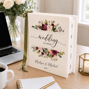 May include: A cream-colored wedding planner binder with a green ribbon bookmark. The cover features floral illustrations in burgundy, pink, and green, with the text "wedding PLANNER", "Madison & Michael", and "2026" printed in black script and serif fonts.