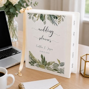 May include: A white three-ring wedding planner binder featuring a botanical green leaf design, the text "wedding planner", "Caitlin & Jason", and "JUNE 25, 2022". It includes a gold ribbon bookmark and white tabbed dividers labeled Timeline, Budget, Attire, Invitations, and Registry.
