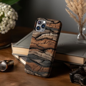 May include: A smartphone case with a wood grain design. The case features a pattern of brown, black, and tan colors, resembling the texture of tree bark. The case is on top of a stack of books.