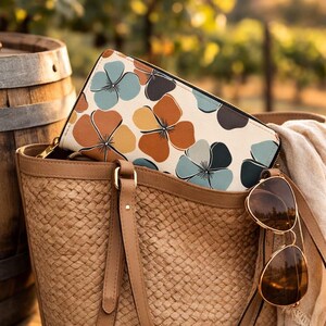 Könnte beinhalten: Eine gewebte Stroh-Tragetasche mit einer Geldbörse mit Blumenmuster im Inneren. Die Geldbörse hat einen cremefarbenen Hintergrund mit blauen, braunen und orangefarbenen Blumendesigns. Sonnenbrillen hängen am Griff der Tasche. Ein Weinglas und Trauben stehen auf einem Holzfass.